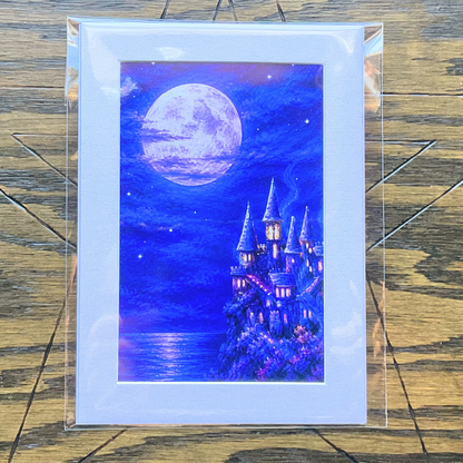 A matted art print depicting a whimsical purple and blue castle under a large glowing moon, mounted in a clear protective sleeve and displayed on a rustic wooden background.