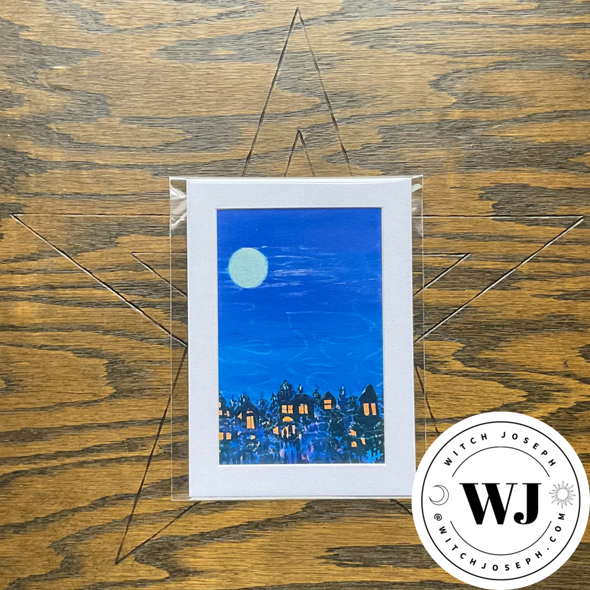 A vertical art print featuring a blue night sky with a pale moon and a row of glowing houses, displayed on a wooden surface with a clear acrylic stand. A circular 'Witch Joseph' brand logo is in the bottom right corner.