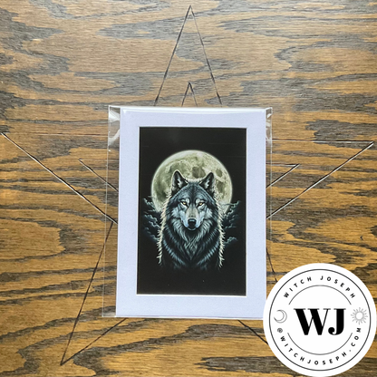 A square art print featuring a detailed illustration of a wolf in front of a full moon, presented in a clear protective sleeve in a white mat. The print is displayed flat on a rustic wooden surface with a black pentagram outline drawn behind it…the Witch Joseph logo is in the bottom right hand corner