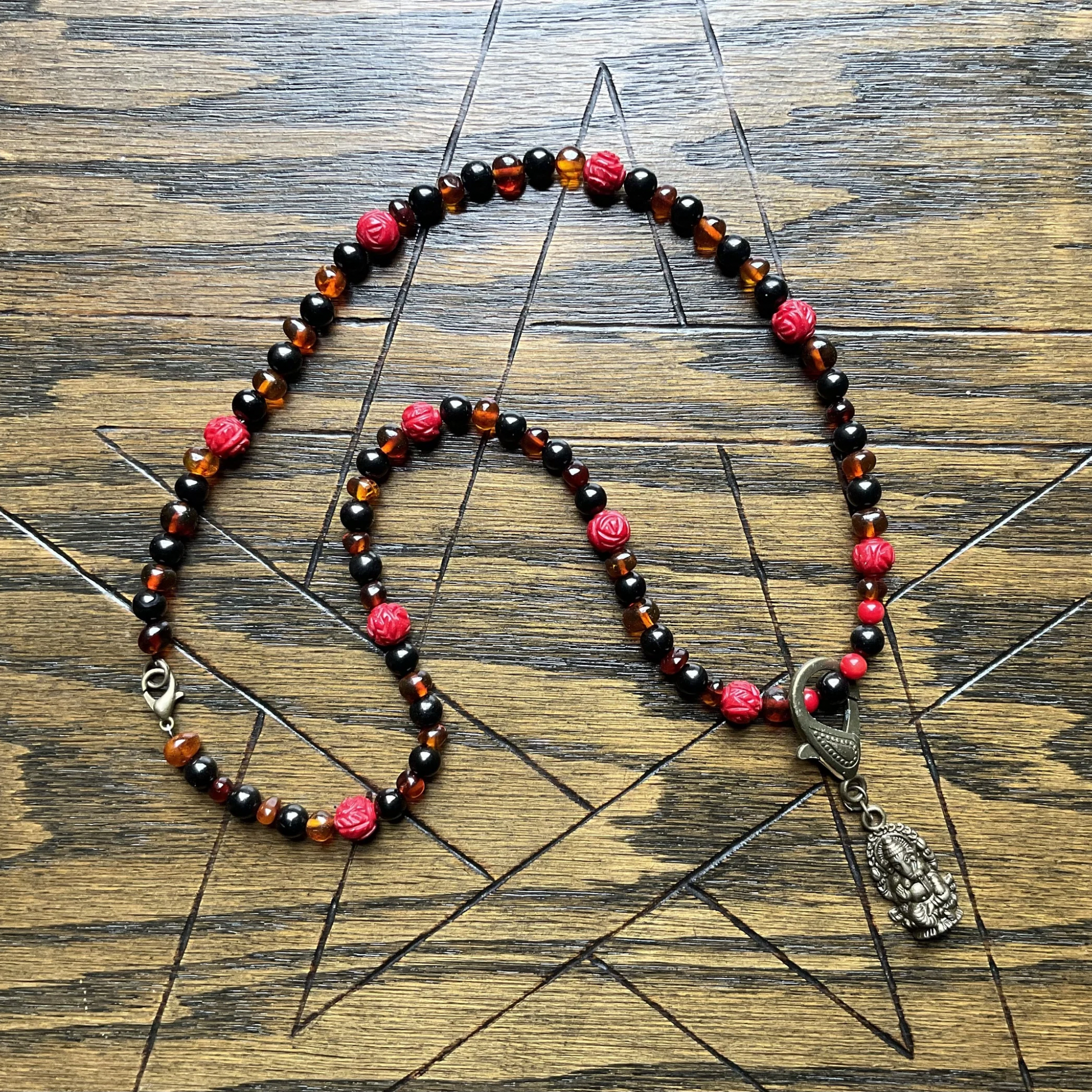 Necklace with amber jet and coral beads and a bronze tone Ganesha pendant displayed on a wooden surface with a Pentagram carved into it