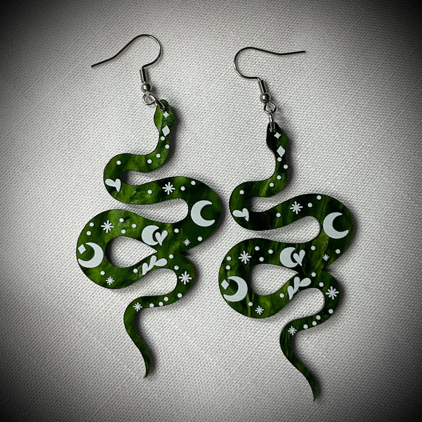 Green snake-shaped earrings with white moon patterns displayed on a linen background