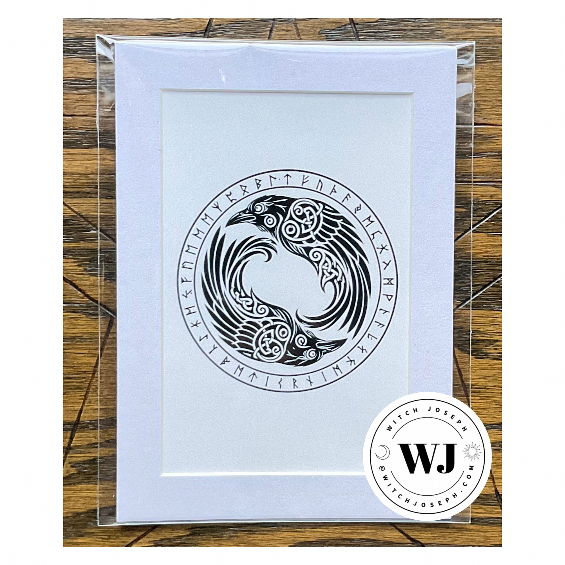 An art print in white mat featuring a black Norse style raven design with runic symbols, enclosed in a clear plastic sleeve and photographed on a wood surface. A circular 'Witch Joseph' logo watermark is visible in the lower right corner.