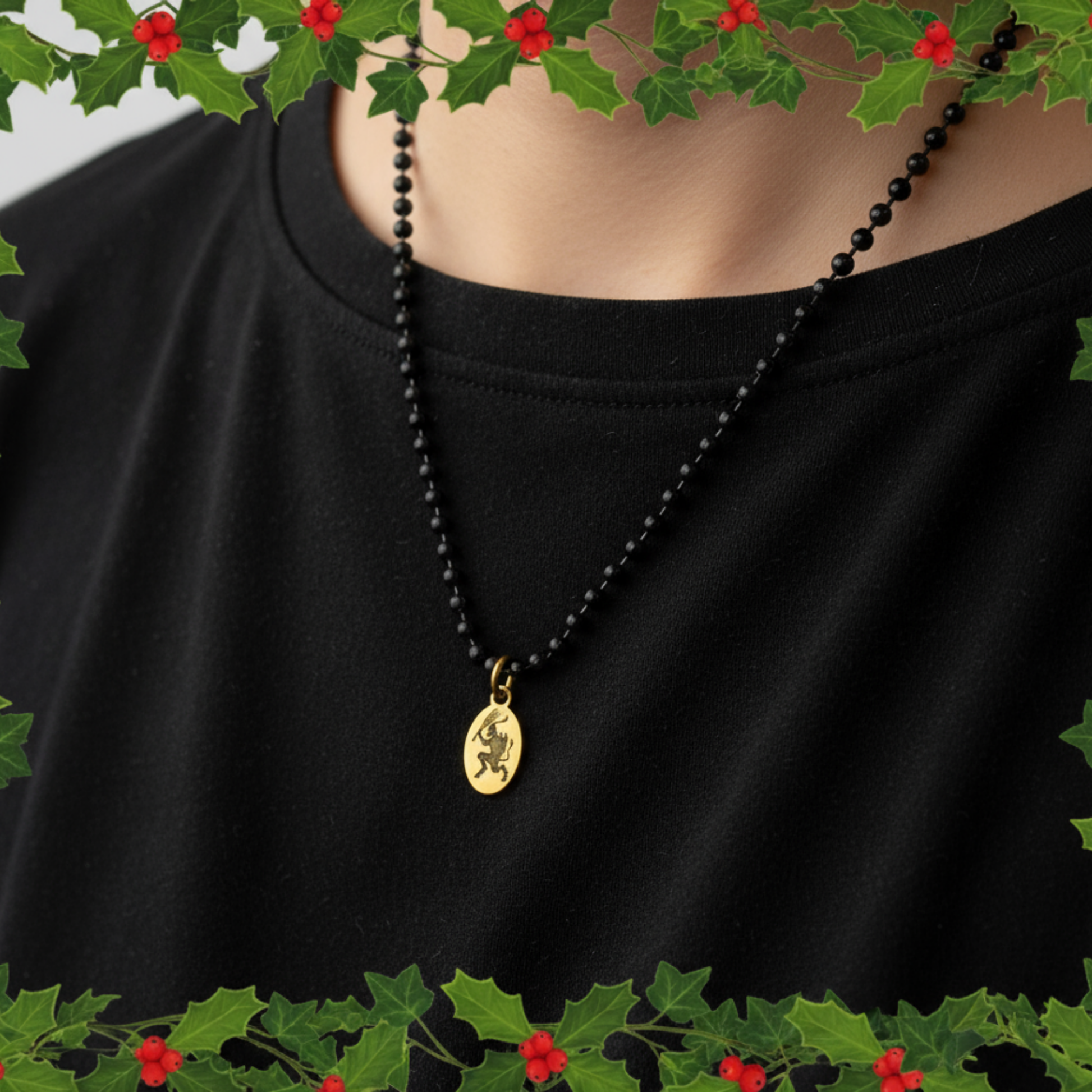 Black ball chain necklace with a brass Krampus pendant on a black shirt, surrounded by holly leaves and berries