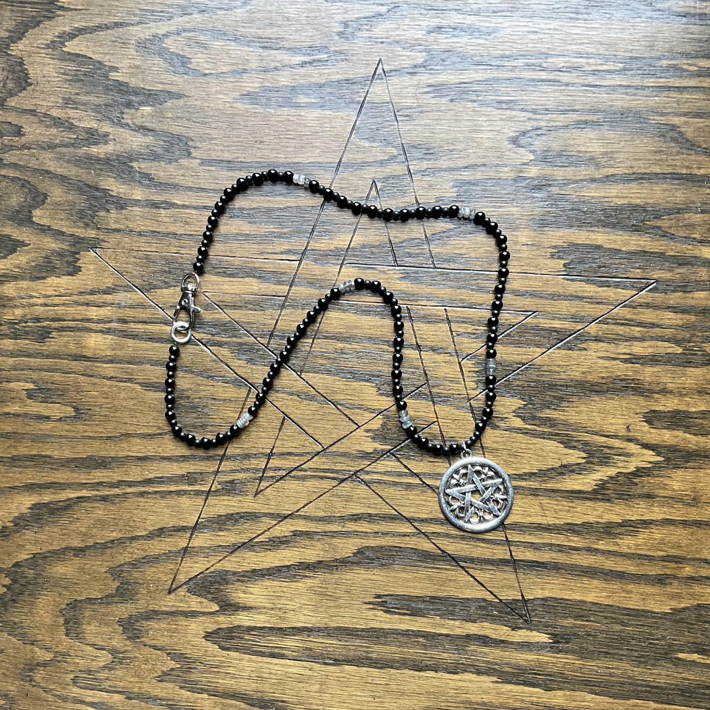 Black beaded Necklace with labradorite chips and with a silver pentacle pendant displayed on a wooden surface with a pentagram etched into it