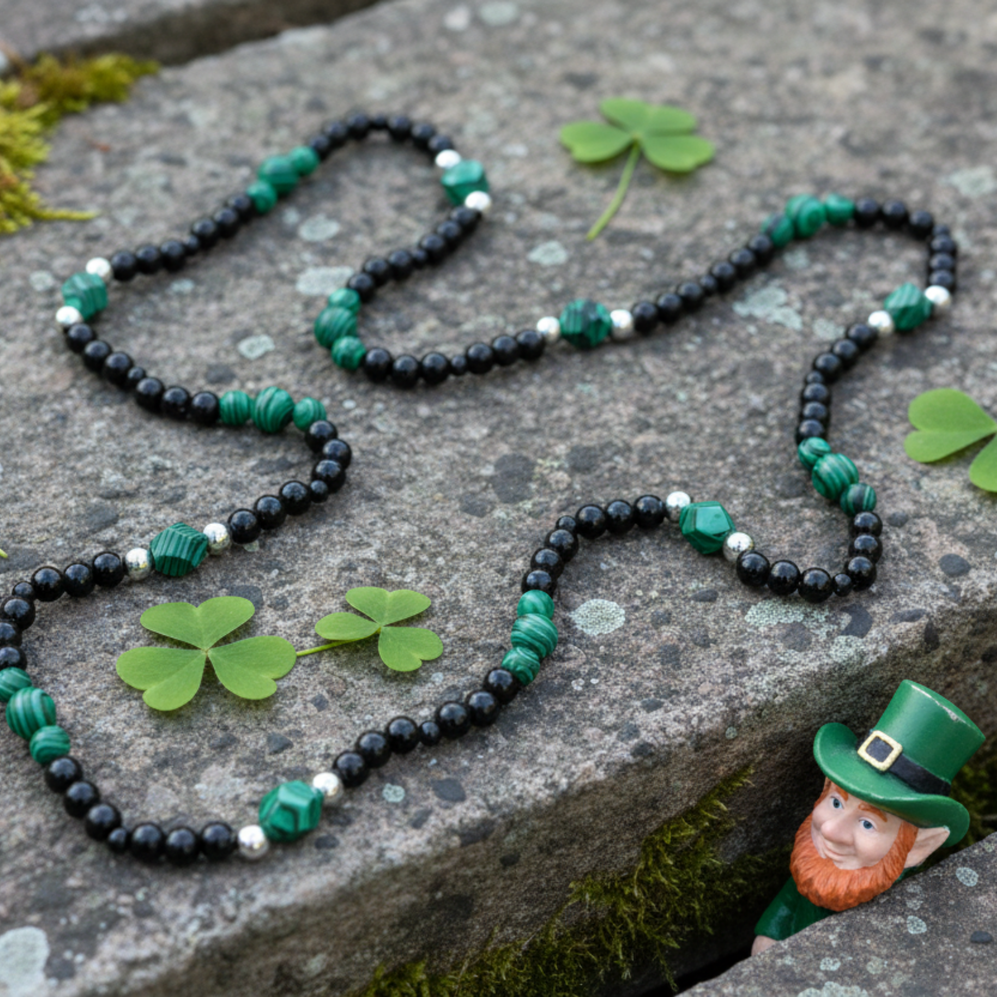 A black beaded necklace with malachite and steel bead accents…displayed on a rustic stone with moss and shamrocks…a small leprechaun is peeking out rom the stone