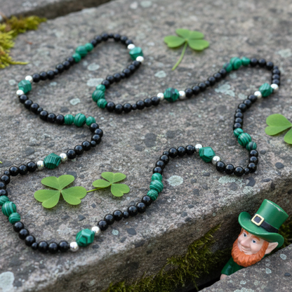 A black beaded necklace with malachite and steel bead accents…displayed on a rustic stone with moss and shamrocks…a small leprechaun is peeking out rom the stone