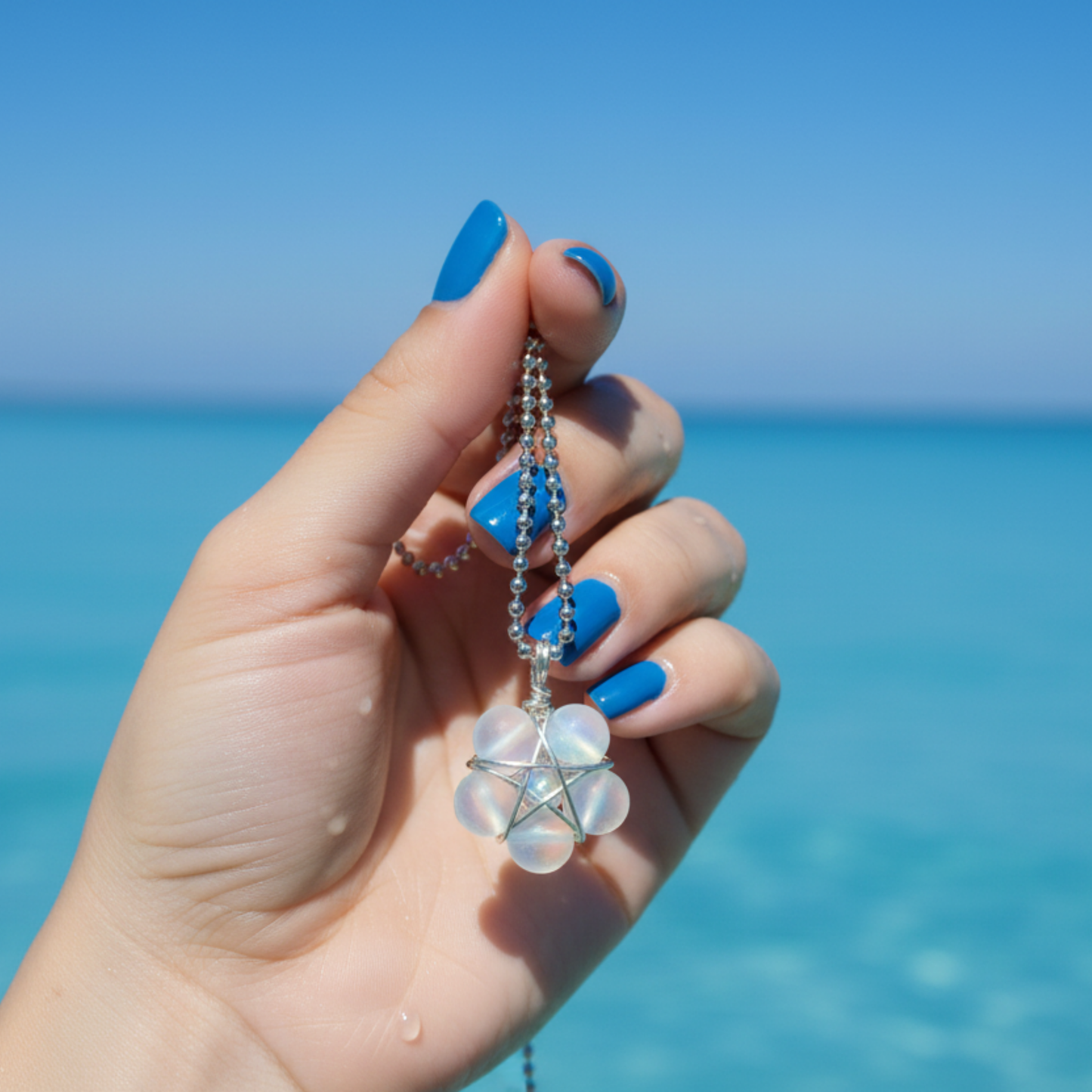 Feminine Hand holding a ball chain necklace with a mermaid bead wire wrap pentagram pendant against a clear blue sky and ocean background
