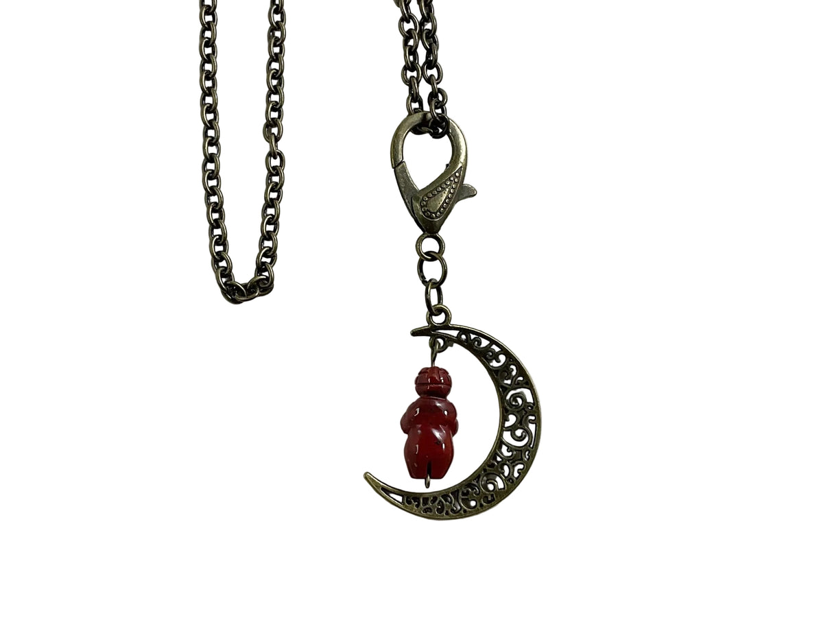 A bronze-toned metal necklace featuring a crescent moon pendant with an ornate filigree design, a small red jasper Goddess bead charm, and a lobster clasp on a white background.