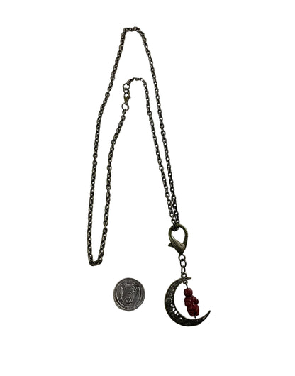 A gothic-style necklace with a dark metal chain, a crescent moon pendant, and small red Goddess bead, shown next to a coin for scale on a plain white background.