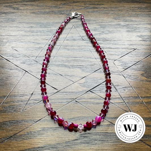 Pink Choker with pink agates and pink crystals beaded together shown on a rustic wood surface with the Witch Joseph Logo