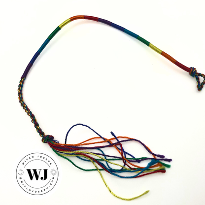 A rainbow braided cord bracelet with a tassel, shown on a plain white background. The product features multiple colors in a rainbow pattern (red, orange, yellow, green, blue, purple) and is finished with a braided closure and a long tassel…shown with the Witch Joseph Logo