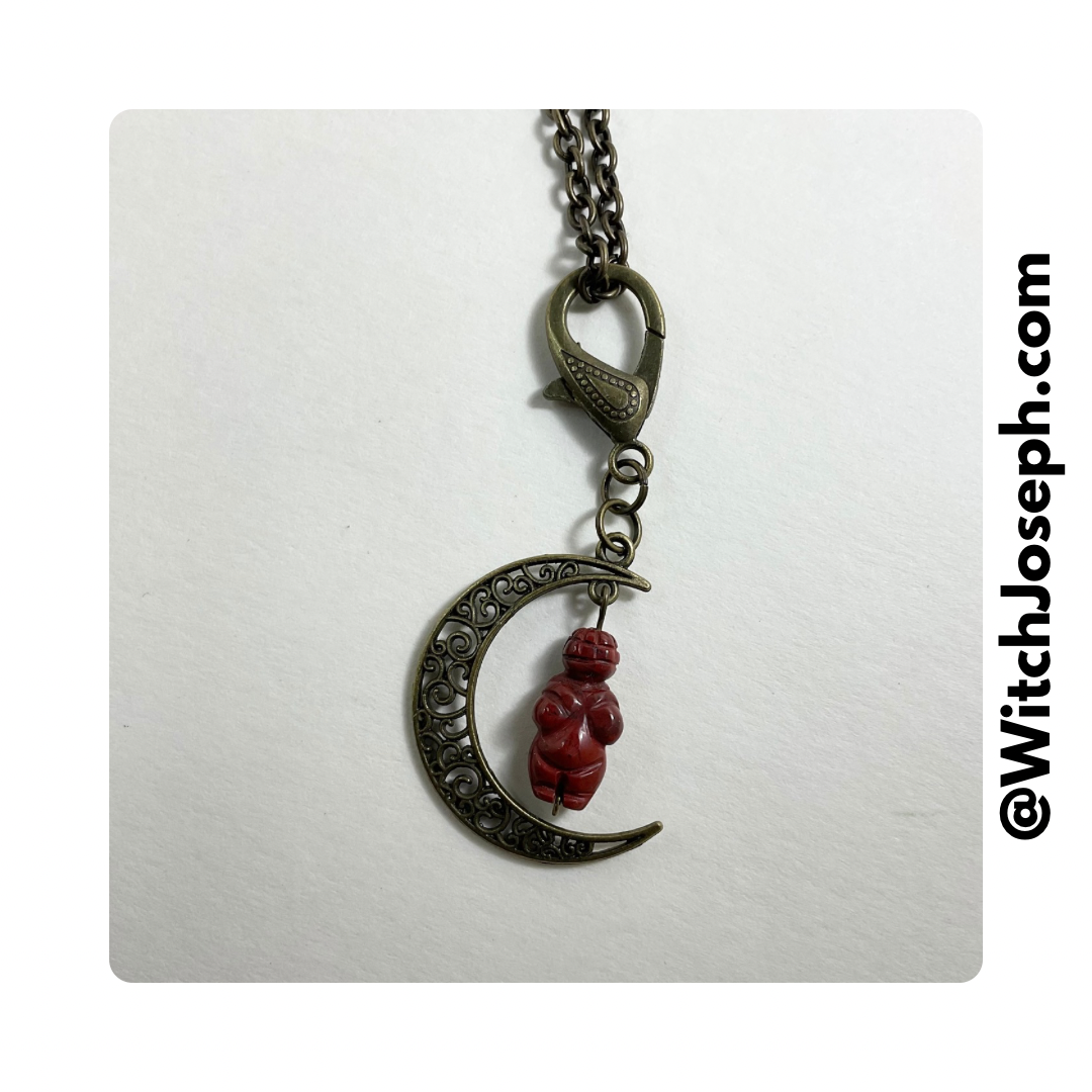 A bronze-toned crescent moon with a red jasper bead pendant, attached to a decorative bronze-toned lobster claw chain.