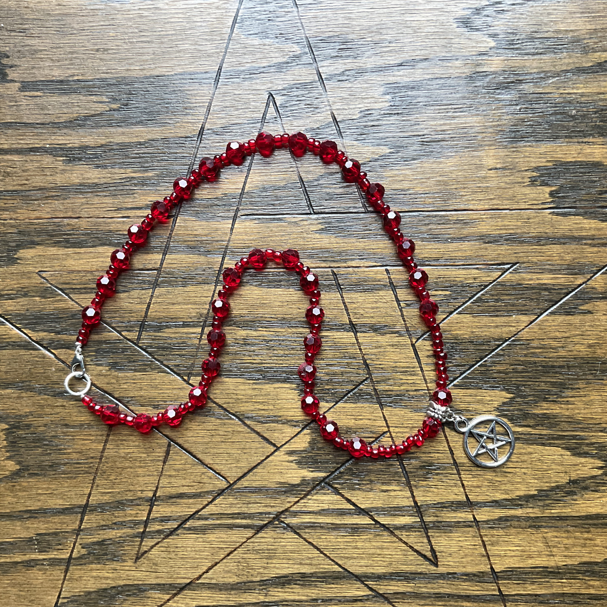 Red crystal beaded necklace with a silver tone Pentacle charm displayed on a wooden surface with a pentagram etched in it