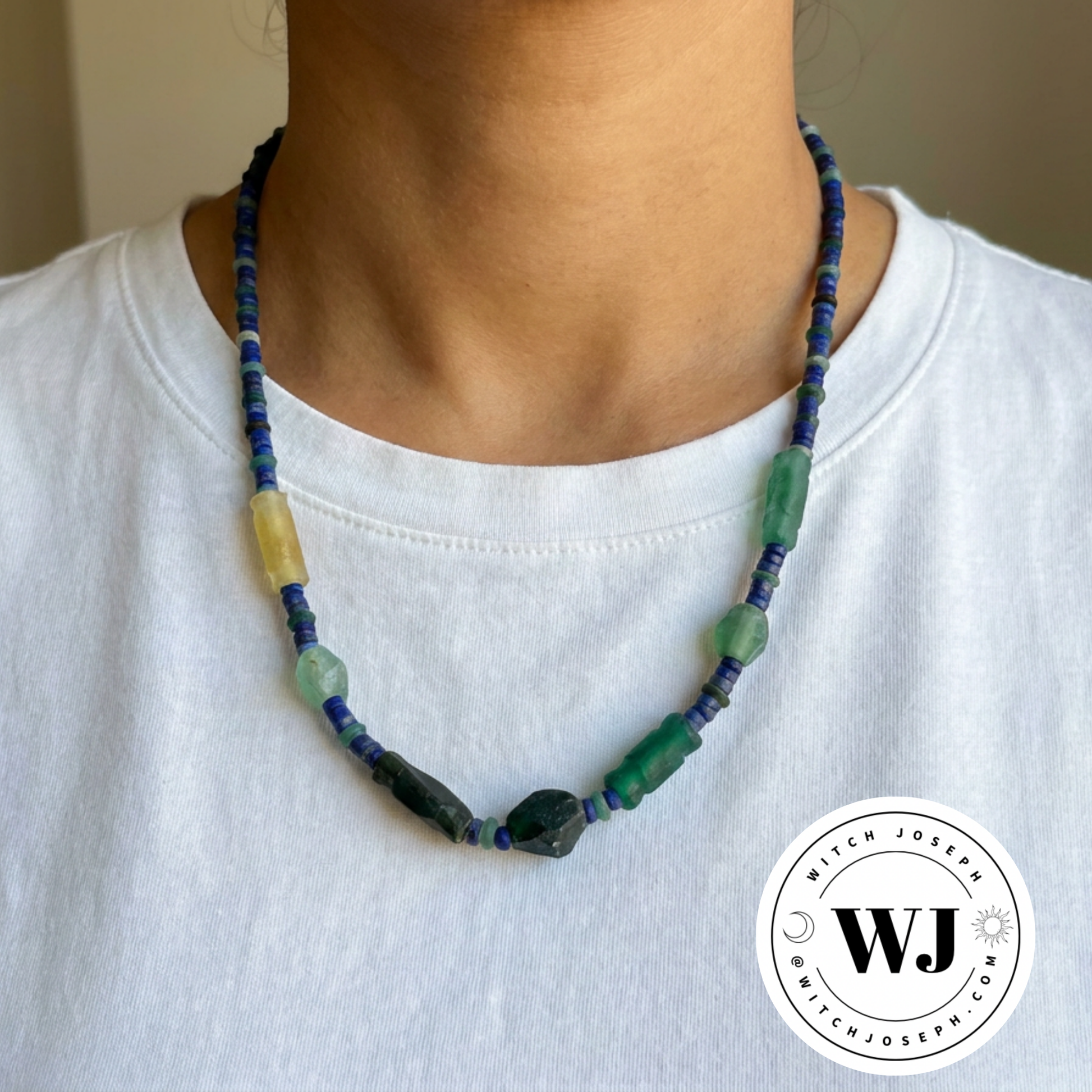 Roman Glass Necklace with blue and green beads and lapis lazuli worn by a model on a white shirt, with the Witch Joseph Logo