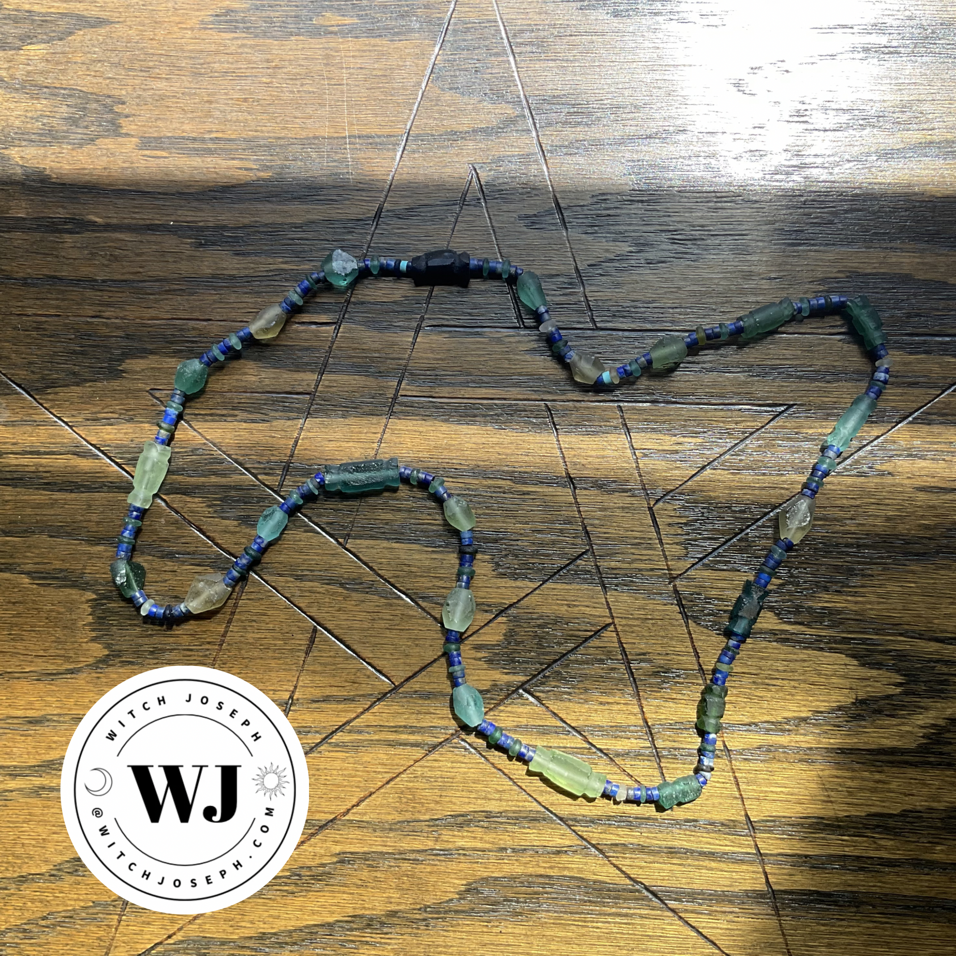 Ancient Roman glass beads in green, yellow and blue with lapis lazuli beaded necklace displayed on a wooden plane with a pentagram etched into it with the Witch Joseph logo