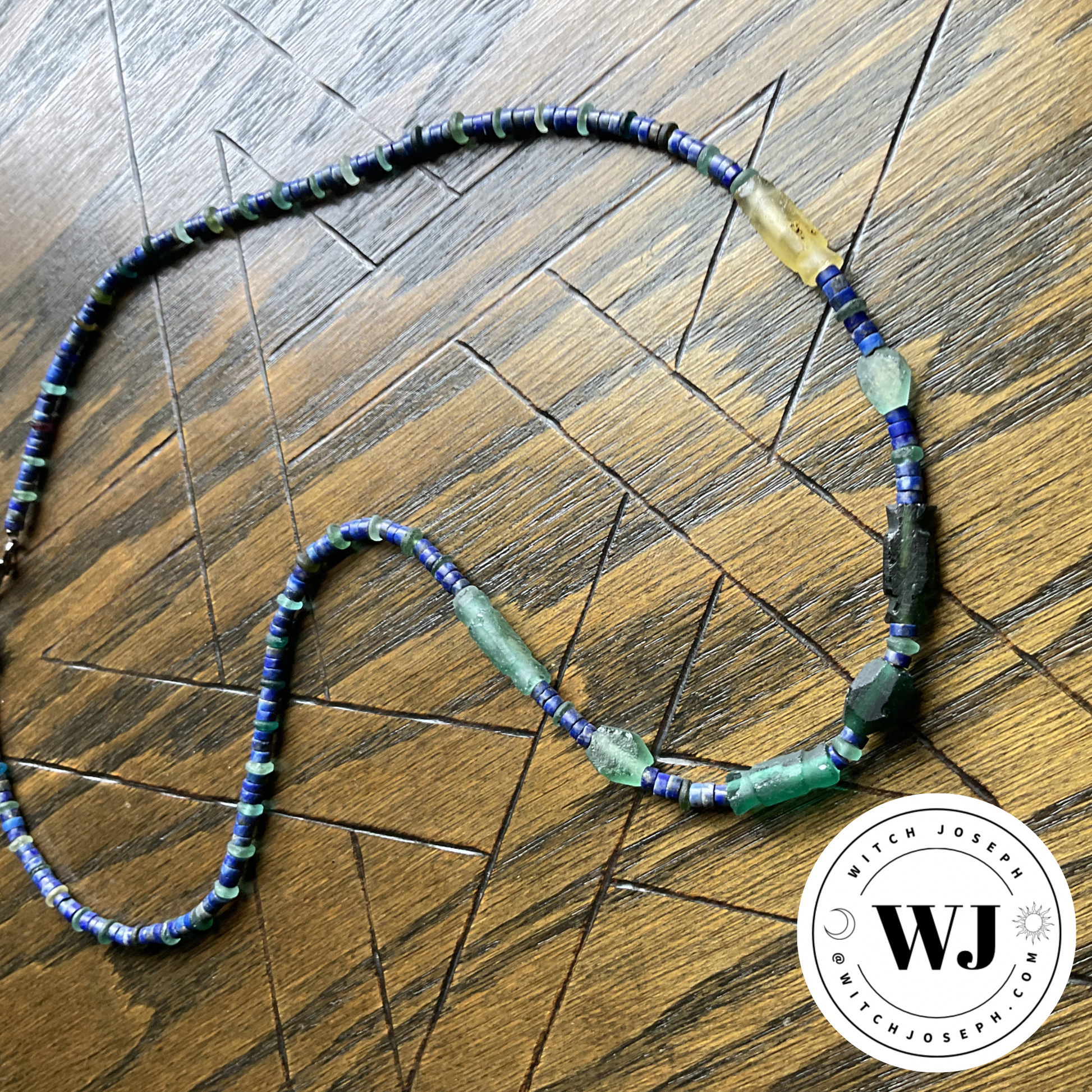 Beaded necklace with Roman glass and lapis lazuli blue and green beads displayed on a wooden surface with an etched pentagram featuring the Witch Joseph logo