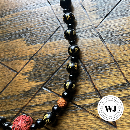 A close-up of a beaded necklace laid diagonally on a dark wood surface. The necklace features glossy black beads with gold Tibetan/Sanskrit script, a textured brown rudraksha seed bead with two smaller ones…displayed on a wooden surface with the Witch Joseph logo in the right lower corner.