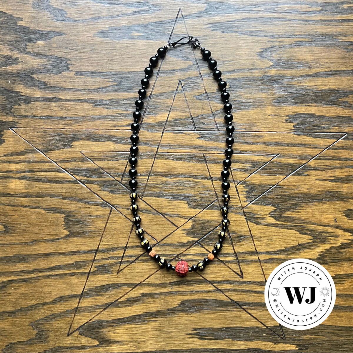 A beaded necklace with black beads and a central red textured bead, displayed on a wooden surface with a geometric pentagram engraving. A circular 'Witch Joseph' brand logo is visible in the bottom right corner.