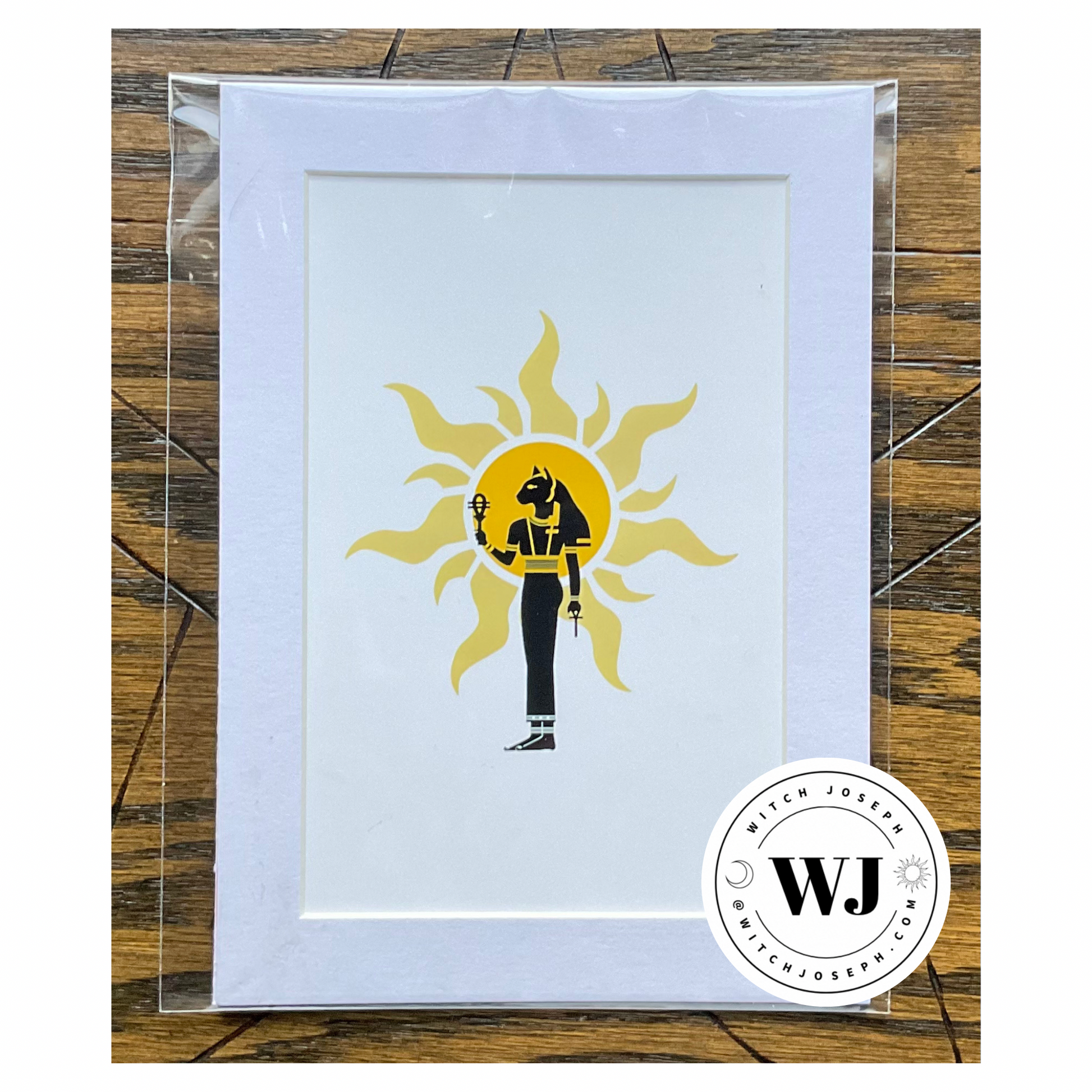 an art print of the Goddess Bastet carrying sistrum and ankh against a solar image with white mat displayed against a wooden background with the Witch Joseph Logo shown in the lower right corner