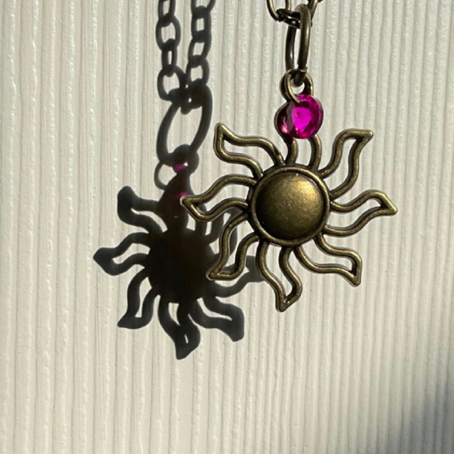 Sun-shaped pendant with a pink gemstone on a chain against a textured beige background