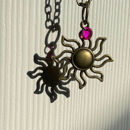 Sun-shaped pendant with a pink gemstone on a chain against a textured beige background