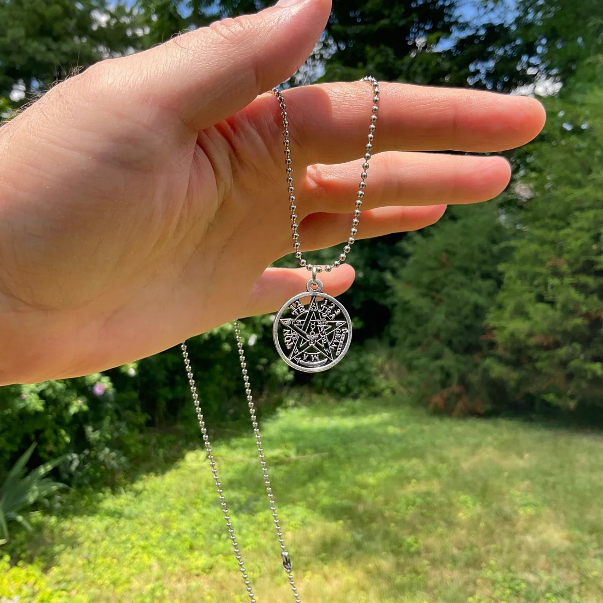 Hand holding a silver necklace with a tetragrammaton pendant against a green garden outdoor background