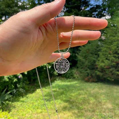 Hand holding a silver necklace with a tetragrammaton pendant against a green garden outdoor background