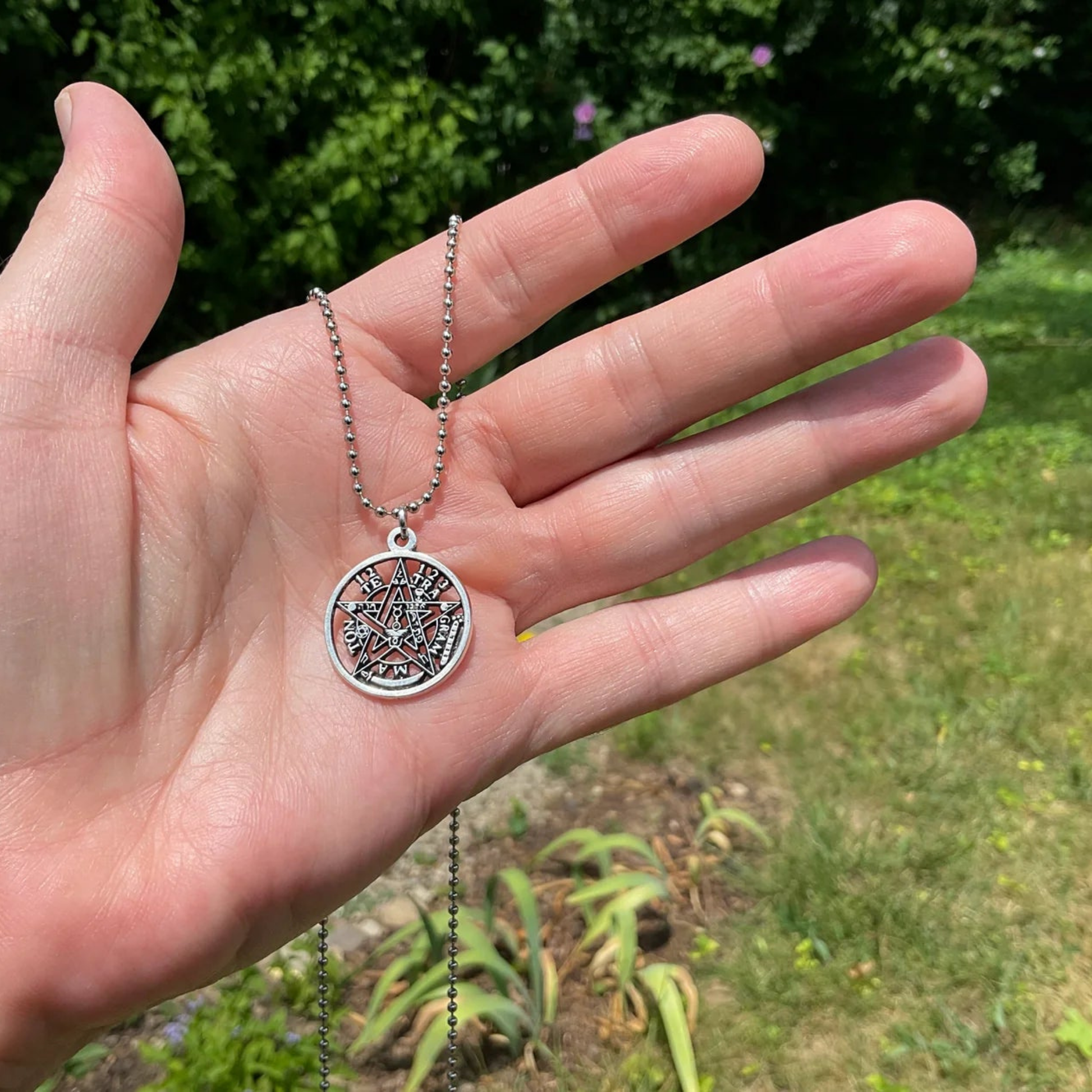Hand holding a silver tone necklace with a circular Pentacle Tetragrammaton pendant against a green garden outdoor background