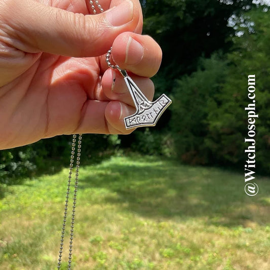 A hand holding a silver Thor's Hammer pendant necklace outdoors with a blurred green grassy background. The pendant features engraved runes.  Also visible is the Witch Joseph URL.