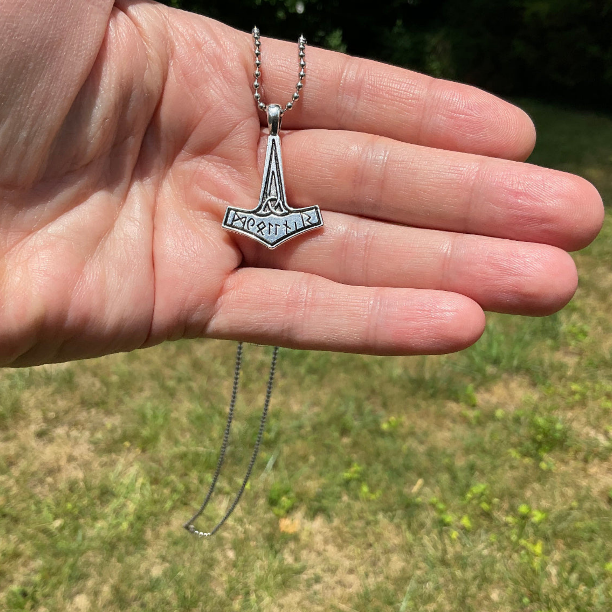 Hand holding a silver tone necklace with a Thor's hammer pendant against a grassy background.