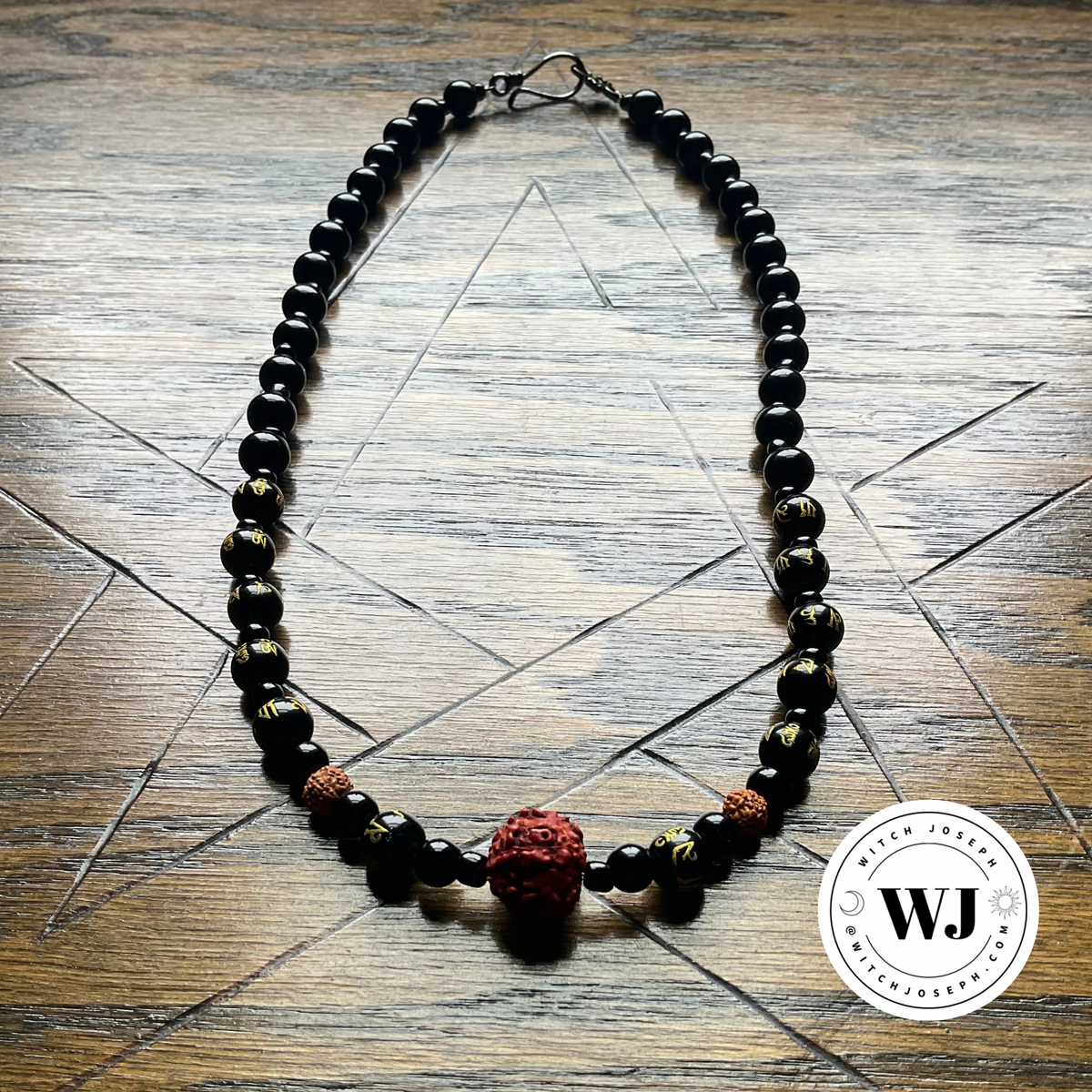 A beaded necklace featuring glossy black beads, two small brown beads with gold patterns, and a prominent textured red rudraksha pendant, displayed on a wooden surface with a pentagram geometric star design and the Witch Joseph logo