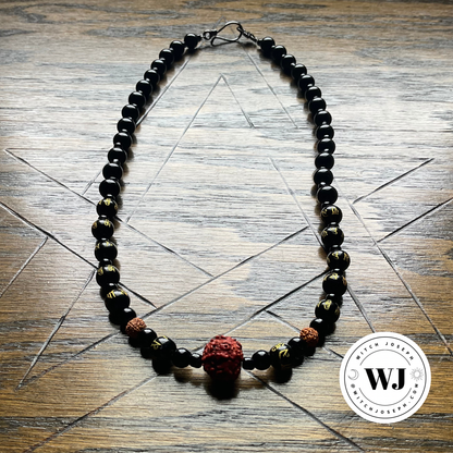 A beaded necklace featuring glossy black beads, two small brown beads with gold patterns, and a prominent textured red rudraksha pendant, displayed on a wooden surface with a pentagram geometric star design and the Witch Joseph logo
