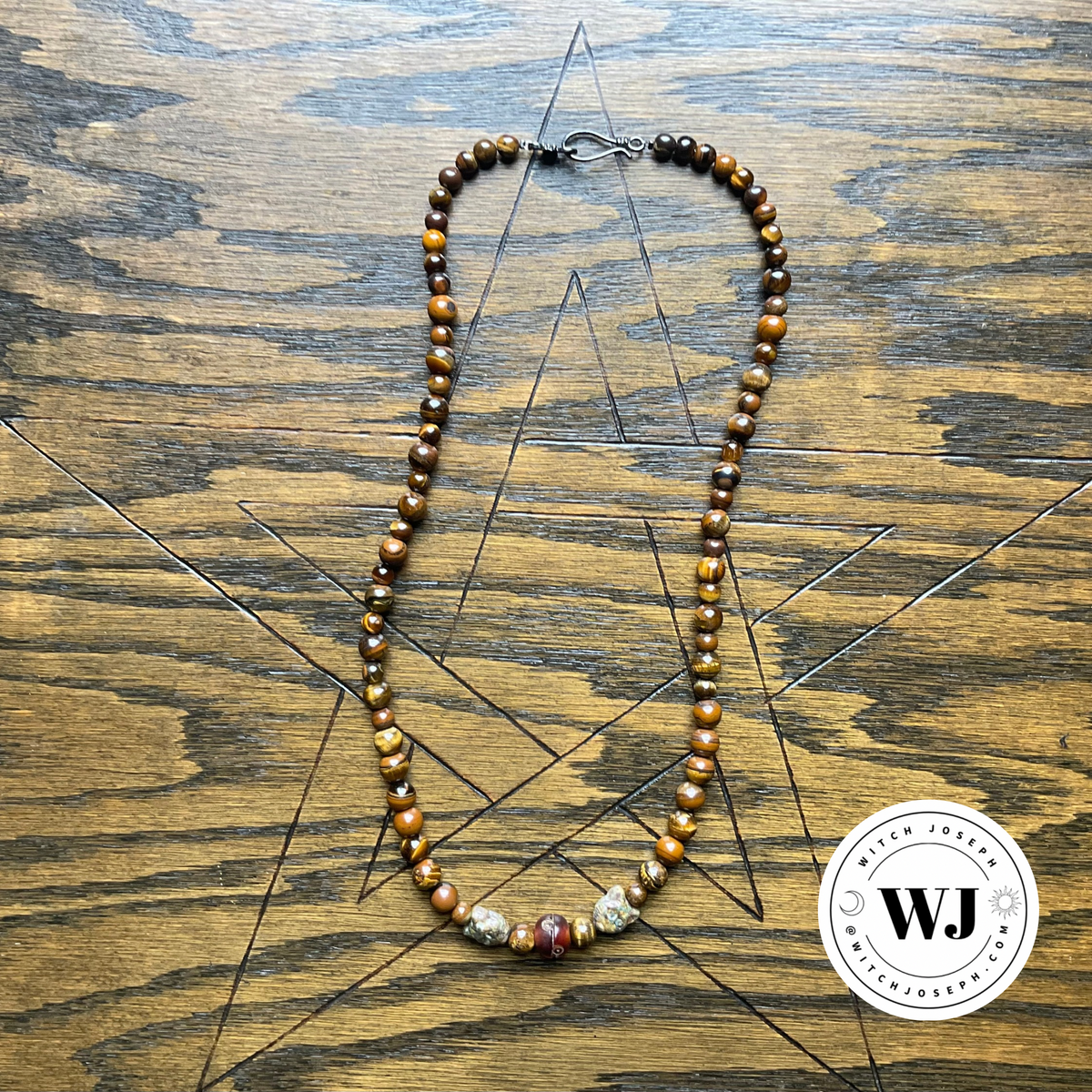 A beaded necklace made of polished brown and gold tiger's eye beads with two cat face Czech beads and a carved accent resin bead, displayed on a wooden surface with a geometric star design. A circular 'Witch Joseph' logo is in the bottom right corner
