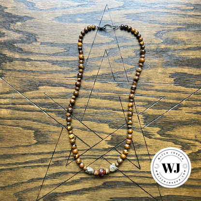 A beaded necklace made of polished brown and gold tiger's eye beads with two cat face Czech beads and a carved accent resin bead, displayed on a wooden surface with a geometric star design. A circular 'Witch Joseph' logo is in the bottom right corner