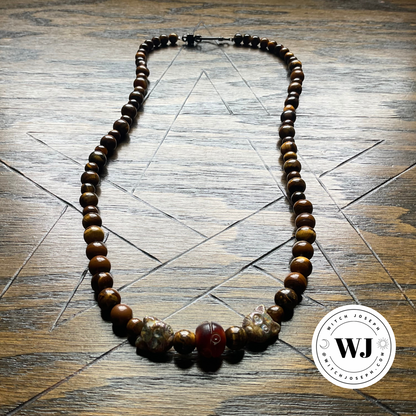 A beaded necklace made of polished brown tiger's eye beads, featuring a central cluster of carved white and reddish beads, displayed on a wooden surface with geometric pentagram line drawings. A circular 'Witch Joseph' logo watermark is visible in the bottom right corner.
