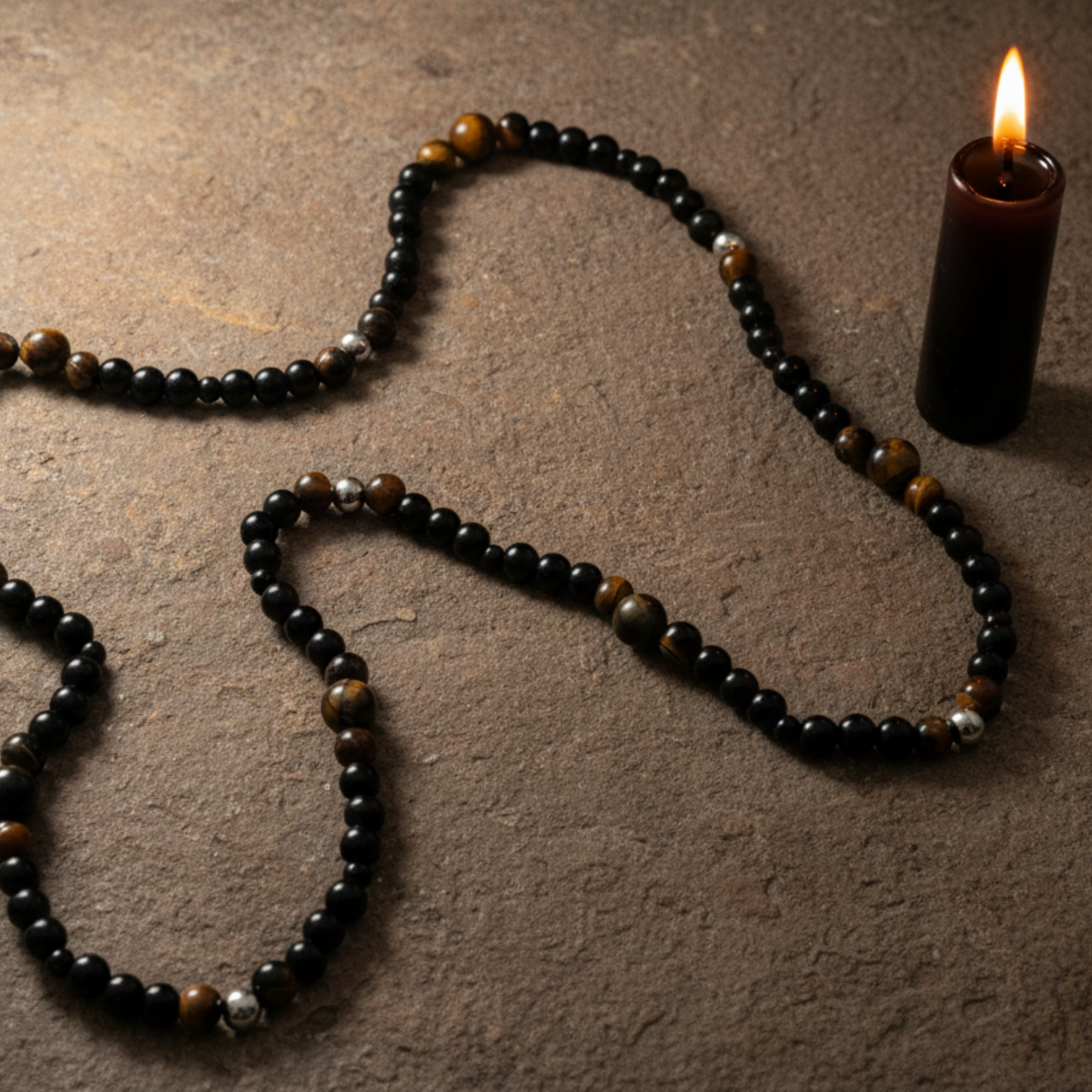 Tigers Eye necklace with black and steel bead accents sitting on a rustic stone next to a lit black candle