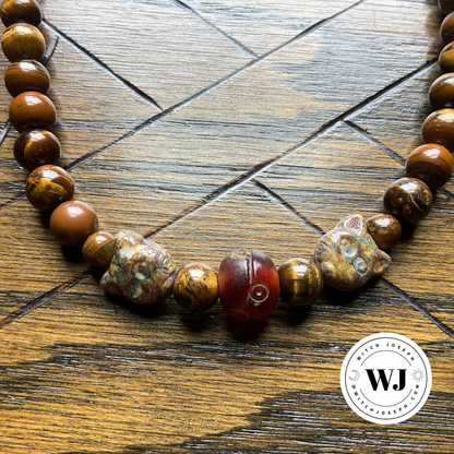A beaded necklace featuring polished brown tiger's eye beads, two Czech Glass Cat Face beads, and a central carved resin bead with detail, displayed on a dark wood background. A circular 'Witch Joseph' logo is visible in the bottom right corner.