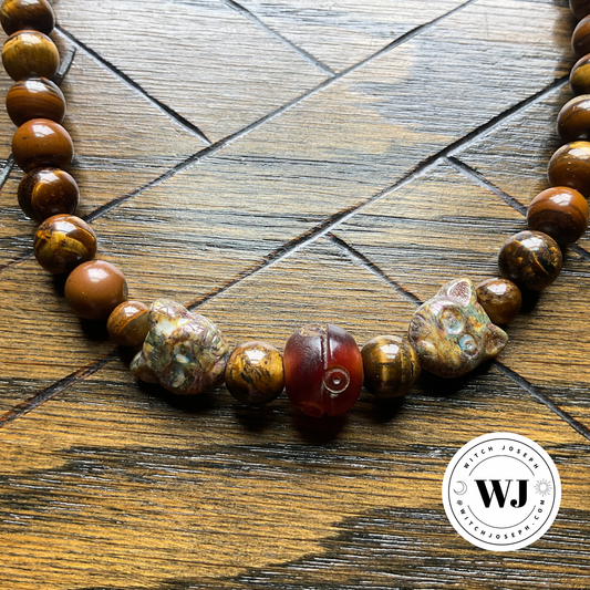 A beaded necklace featuring polished brown tiger's eye beads, two Czech Glass Cat Face beads, and a central carved resin bead with detail, displayed on a dark wood background. A circular 'Witch Joseph' logo is visible in the bottom right corner.