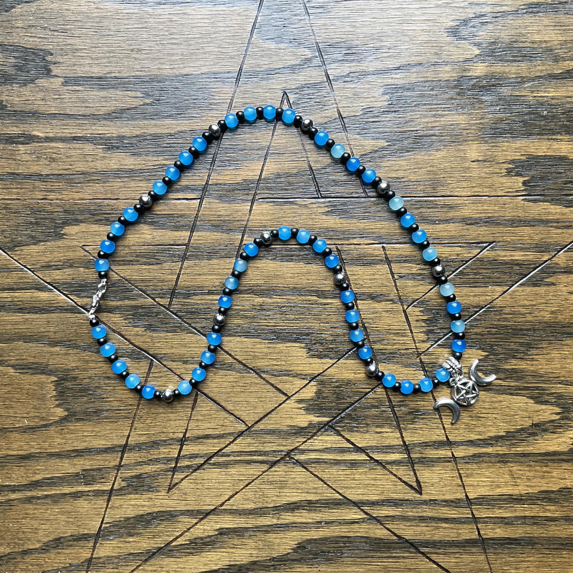 Necklace with blue and black beads with a silver tone triple goddess charm displayed on a wooden surface with a pentagram etched into it