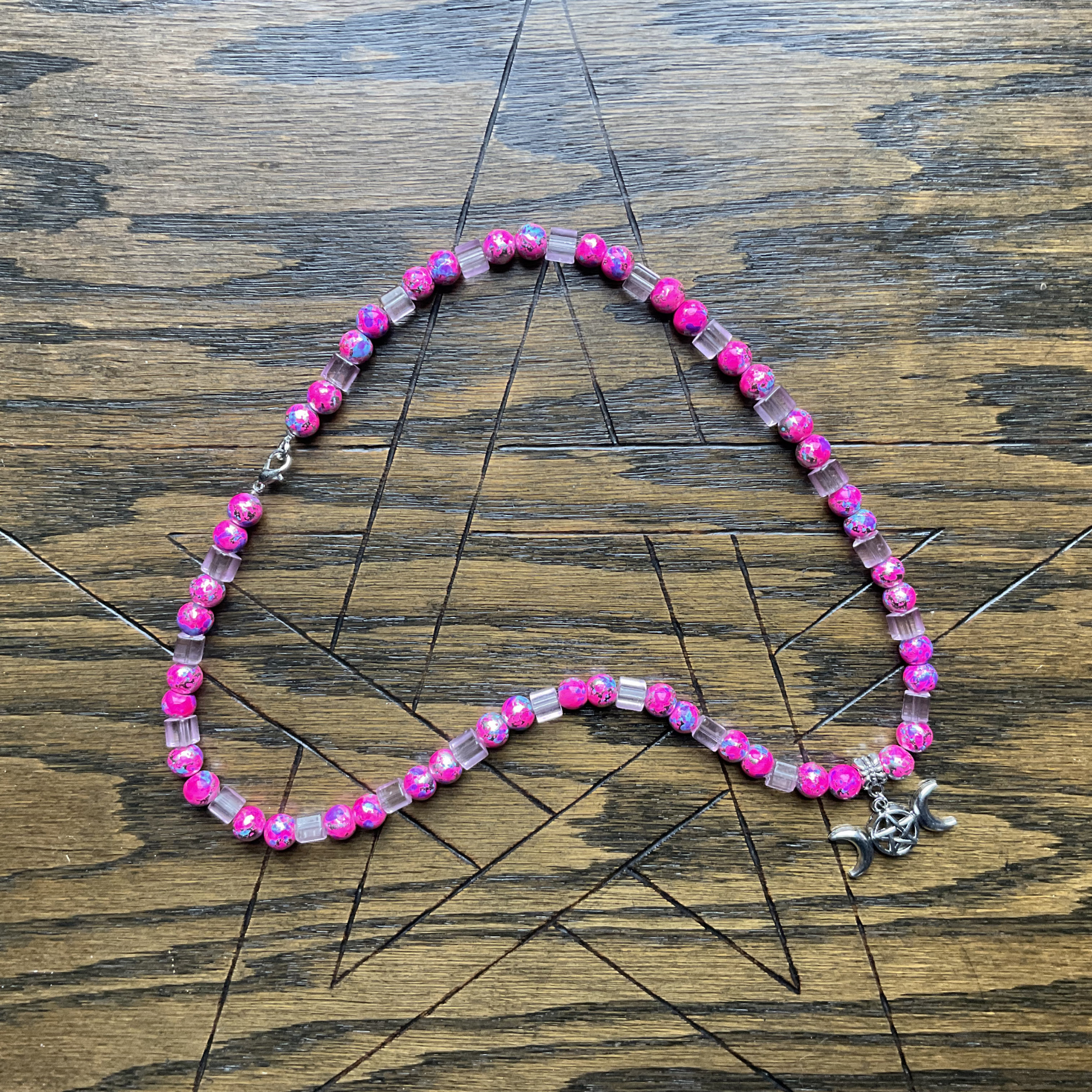 Beaded necklace with pink and lite pink beads with a silver tone Triple Goddess Pentacle Pendant displayed on a wooden surface with a Pentagram etched into it