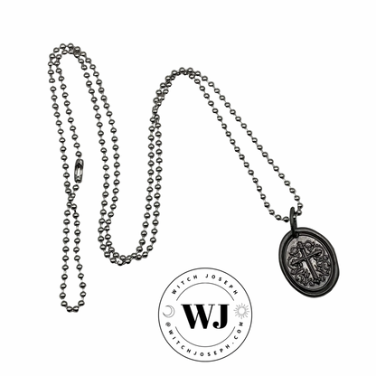 Gunmetal tone oval sealing wax style Cross charm on a silver tone ball chain necklace displayed on a white background, featuring the Witch Joseph logo