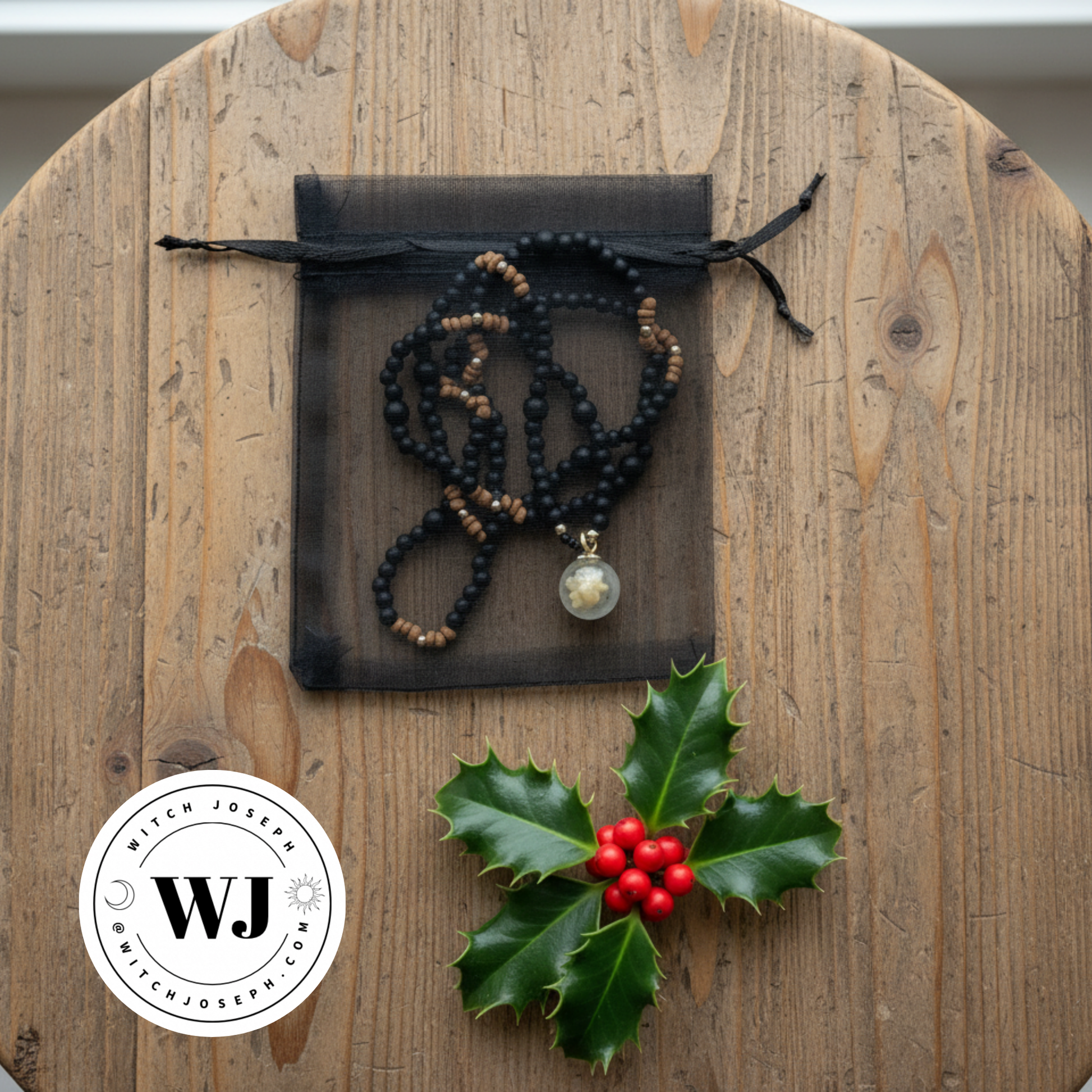 Three kings magi necklace shown on it’s black organza gift bag on a wood table with a piece of holly and the Witch Joseph logo
