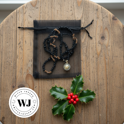 Three kings magi necklace shown on it’s black organza gift bag on a wood table with a piece of holly and the Witch Joseph logo