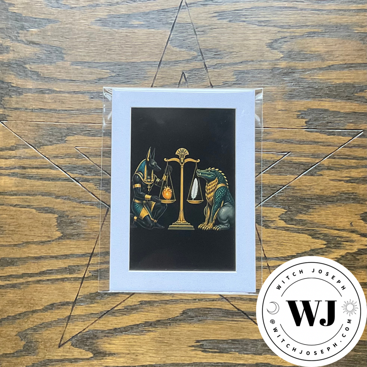 An art print featuring a black background with a central illustration of a golden scale of justice, flanked by Ammit on the right and Anubis on the left. The print has a white border and is shown inside a clear protective sleeve displayed on a rustic wooden background with a star design with the witch joseph logo in the lower right corner.