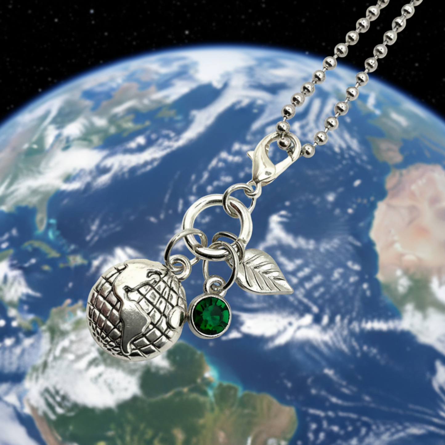Silver tone Earth Day necklace with globe charm and a silver tone leaf charm and a green crystal charm…hanging against a backdrop of Earth in space.