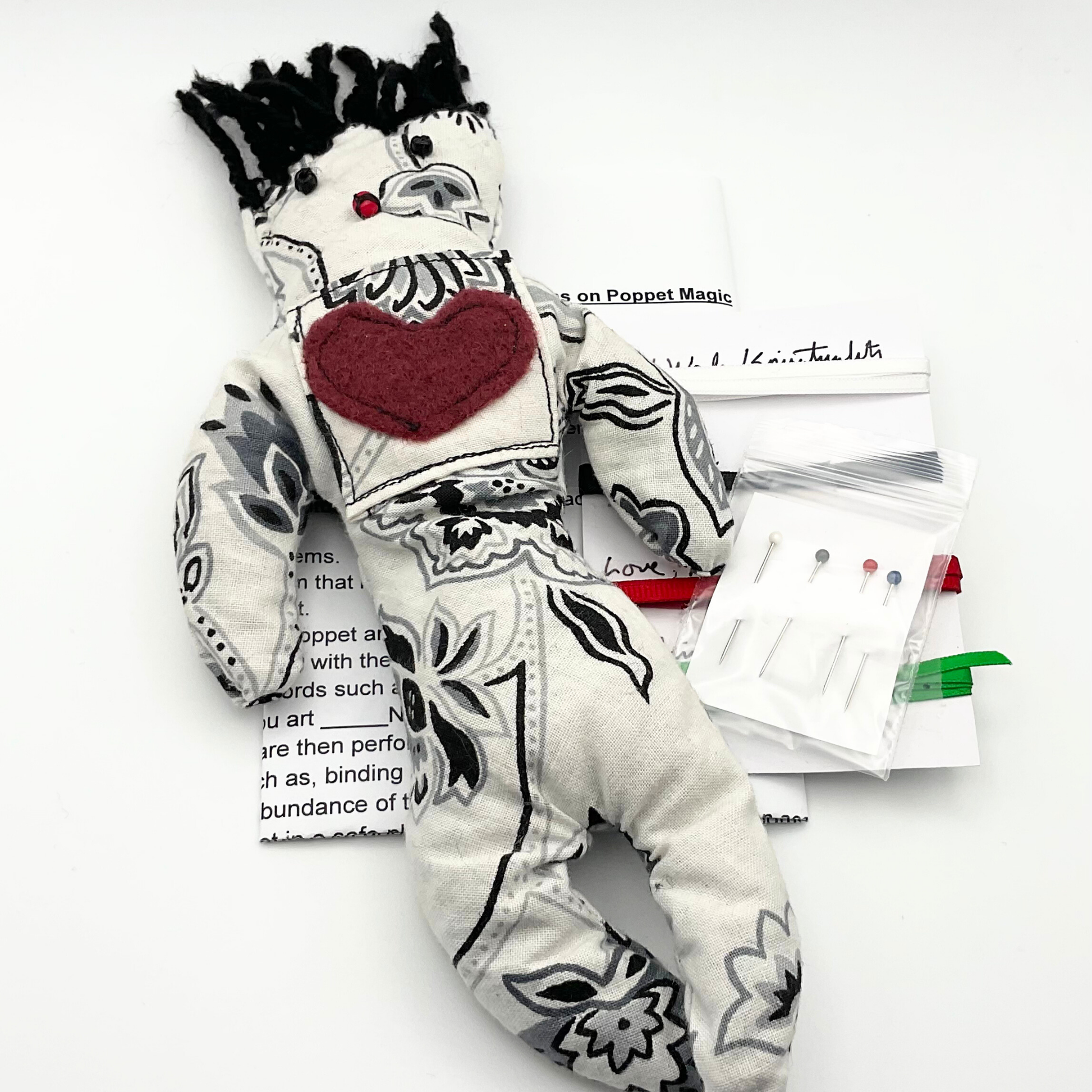 Stuffed poppet with black and white patterns and a red heart, next to a set of needles and ribbons on a white background.